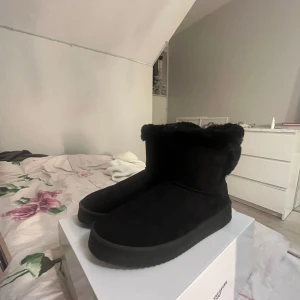 Svarta fluffiga boots i mocka uggs  - Mysiga svarta boots med fluffigt foder och ovandel i mocka. Skorna har rund tå och platt sula, perfekta för kalla dagar. De är helt svarta och har en enkel, stilren design som passar till det mesta.