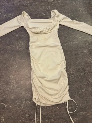 Vit långärmad omlottklänning - Snygg vit klänning med långa ärmar och omlottdetaljer. Klänningen har rynkade sidor med snörning nertill och en fyrkantig urringning. Perfekt för dig som gillar en stilren look med lite extra detaljer. Har en liten  smink fläck som nog går att tvätta bort .