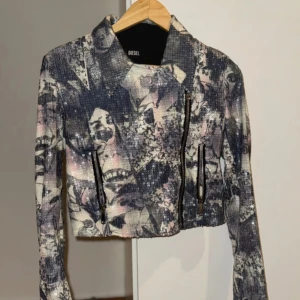 Mönstrad croppad jacka från Diesel - Unik croppad jacka från Diesel med collage-liknande mönster i svart, grått, vitt och inslag av rosa. Jackan har långa ärmar, coola dragkedjor framtill och på ärmsluten samt en klassisk krage. Materialet har en lätt glittrig finish som ger extra edge.