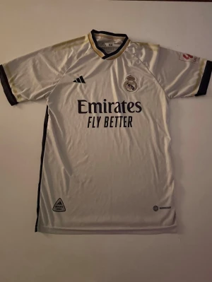 Real Madrid Bellingham matchtröja - Säljer en officiell Real Madrid matchtröja från Adidas med Bellingham och nummer 5 på ryggen. Tröjan är vit med svarta och guldiga detaljer, har klubbmärke och sponsortryck på bröstet samt Aeroready-material för extra komfort. Perfekt för dig som älskar fotboll och Real Madrid.