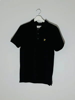 Svart piké från Lyle & Scott - Svart pikétröja från Lyle & Scott med klassisk krage och tre knappar framtill. På bröstet finns den ikoniska gula fågeln broderad. Materialet är bomull och modellen har korta ärmar. Perfekt för en clean och stilren look.