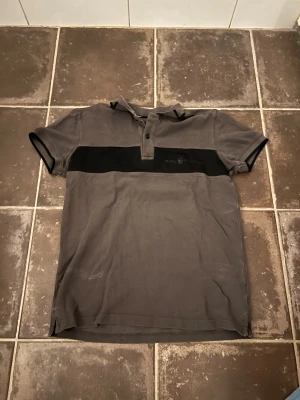 Sail racing pike  - Säljer en grå t-shirt från sail racing med svart horisontellt band över bröstet och diskret logga. T-shirten har korta ärmar, krage med två knappar och är gjord i mjuk bomull. Perfekt för dig som gillar stilren och sportig design.