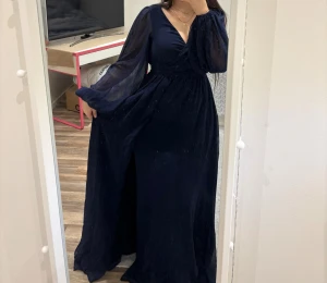 Mörkblå långklänning med slits - Elegant mörkblå lång balklänning med v-ringning och transparenta, långa ärmar. Klänningen har omlottliv, markerad midja och en snygg slits framtill. Endast använd 1 gång, ord pris 999kr