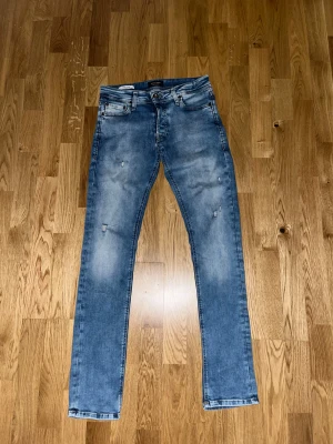 Blå skinny jeans från Jack & Jones - Snygga blå jeans från Jack & Jones med skinny passform och den har, slitna detaljer och brunt läderpatch bak i midjan. Perfekt för dig som gillar en smal siluett och bekväma jeans. Storlek 27/32