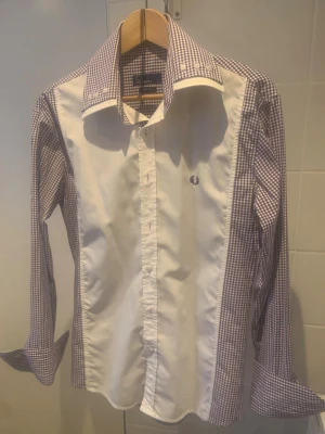 Rutig skjorta från Fred Perry slim fit - Snygg slim fit skjorta från Fred Perry med lila och vit rutigt mönster på ärmar, krage och rygg. Framsidan är vit med broderad logga på bröstet. Klassisk krage med knappar och långa ärmar. Perfekt för dig som gillar stilrena detaljer.