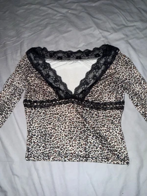 Leopardmönstrad topp med spets - Säljer en snygg leopardmönstrad topp med lång ärm och bred svart spets runt v-ringad halsringning. Toppen har en figurnära passform och coola beige, bruna och svarta toner i mönstret. Perfekt för dig som gillar att sticka ut med djurmönster och detaljer.