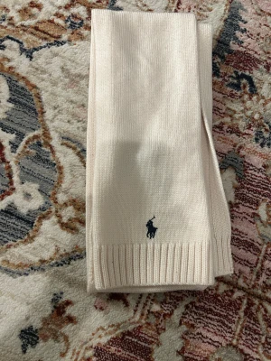 Beige stickad väst från Polo Ralph Lauren - Snygg beige stickad väst från Polo Ralph Lauren med klassisk ribbad kant och den ikoniska mörkblå Polo-loggan broderad framtill. Tillverkad i 100% ull, perfekt för dig som gillar stilrena och tidlösa plagg med premiumkänsla.