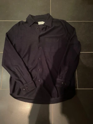 Overshirt från East West - Säljer en marinblå overshirt från East West i storlek M. Klassisk design med krage, bröstficka och knappar hela vägen. Långärmad och tillverkad i mjukt bomullstyg som känns skönt mot huden. Perfekt för dig som gillar stilrena och enkla plagg. Vid fler frågor eller funderingar är det bara att skriva 😊