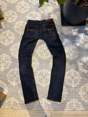 Mörkblå jeans från Nudie Jeans Co W31 L36 - Snygga mörkblå jeans från Nudie Jeans Co med klassiska orange sömmar och ikonisk broderad bakficka. Modellen har smal passform och raka ben, tillverkade i slitstarkt denim. Perfekta för dig som gillar stilrena och tidlösa jeans. Lite lagade i skrevet vilket syns på sista bilden men det är bara att höra dig om du undrar något MVh theo