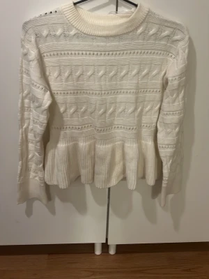 Vit stickad tröja med volang från Gina Tricot - Supersöt vit stickad tröja från Gina Tricot med snygga strukturerade detaljer och volangkant nedtill. Tröjan har rund hals och långa ärmar, perfekt för att ge din outfit en mysig och trendig vibe.