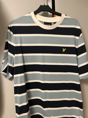 Randig t-shirt från Lyle & Scott - Helt oanvänd snygg t-shirt från Lyle & Scott i storlek M med breda horisontella ränder i marinblått, ljusblått och vitt. Klassisk rund hals med vit kant och gul broderad logga på bröstet. Perfekt för dig som gillar stilrena och sportiga plagg. Säljer pga av att den inte passar. Ordinare pris 600 
