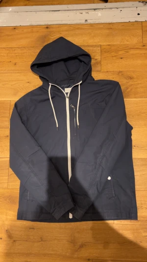 Mörkblå vindjacka från Pier One - Snygg mörkblå vindjacka från Pier One med huva och vita snören. Jackan har vit dragkedja framtill och en enkel, stilren design. Perfekt för blåsiga dagar och enkel att matcha med det mesta. Lätt och smidig att bära med sig.