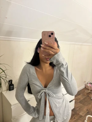 Grå långärmad topp med knyt - Säljer en ljusgrå långärmad topp med djup v-ringning och snygg knytning framtill. Toppen har en slim passform och är tillverkad i mjukt stretchigt material som sitter skönt på kroppen. Perfekt att styla med jeans eller kjol för en trendig look.