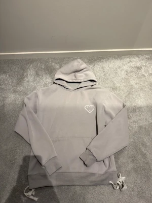 Ljusgrå hoodie från Carsicko med tryck - Säljer en ljusgrå hoodie från Carsicko i storlek S. Hoodien har en stor huva, magficka och ribbade muddar. På bröstet finns ett vitt tryck med ett hjärta och ansikte. Mjuk insida och snörning nedtill. Perfekt för dig som gillar clean streetwear.