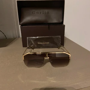 Corlin eyewear glasögon  - Bilderna visar ett par solglasögon från Corlin Eyewear, modell Kelly, med guldiga bågar och bruna tonade glas. Ingen väst syns på bilderna.