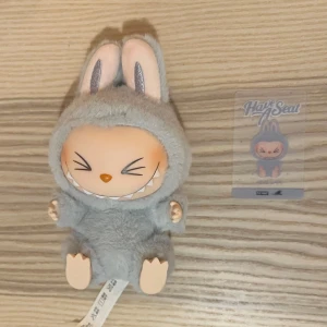 Labubu från Pop Mart - Söt och rolig grå Labubu från Pop Mart med stora öron och ett brett leende. Perfekt som samlarobjekt eller dekoration. Kommer med ett matchande kort. Denna har QR kod på etiketten. Köptes från Popmart i Hong Kong.