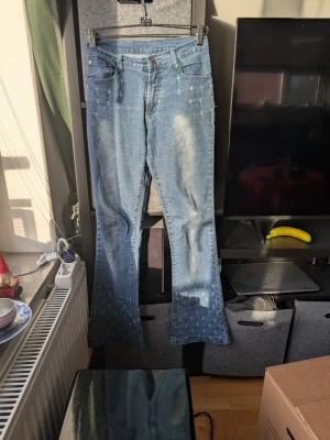 Blå bootcut jeans med broderi Humana - Snygga ljusblå jeans från Humana med bootcut passform och broderade detaljer i form av små kors på fickor och nedre delen av benen. Jeansen har klassisk femficksdesign, knapp och dragkedja framtill samt en ljus tvätt med slitningar.