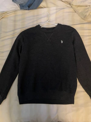 Mörkgrå sweatshirt från Polo Ralph Lauren - Snygg mörkgrå sweatshirt från Polo Ralph Lauren med klassisk rund hals och broderad logga i vitt på bröstet. Tröjan har långa ärmar och ribbade muddar vid ärmslut och nederkant. Perfekt för chill dagar och enkel att matcha med jeans.