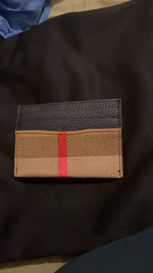 Burberry plånbok med klassiskt mönster - Snygg plånbok från Burberry med klassiskt rutig design i beige och brunt, samt en röd rand i mitten. Övre delen är i svart skinn med plats för kort. Perfekt för dig som vill ha en stilren och ikonisk accessoar i fickan.