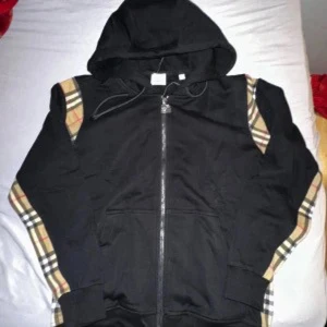 Svart Burberry hoodie med dragkedja - Svart hoodie från Burberry med klassiska rutiga detaljer på axlar och ärmar. Dragkedja framtill, huva med snören och två fickor. Snygg streetstil och skön passform, perfekt för dig som gillar exklusiva märken och vill sticka ut.