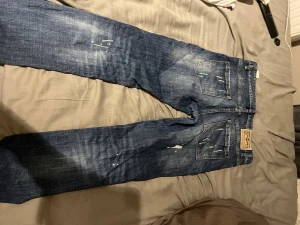 Dsquared2 blå jeans med slitningar, stl 31 - Säljer ett par blå jeans från Dsquared2 i storlek 31. Jeansen har coola slitningar, hål och blekta detaljer samt vita färgstänk för en edgy look. Klassisk femficksmodell med knappgylf och raka ben. Perfekt för dig som gillar streetstyle.