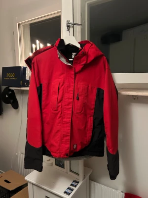 Röd och svart vindjacka från Craft - Säljer en röd och svart vindjacka från Craft med huva, flera fickor med dragkedja och broderad logga på ärmen. Jackan har justerbara ärmslut och meshfoder på insidan. Perfekt för dig som gillar sportig stil och funktionella detaljer.