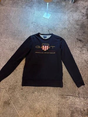 Svart sweatshirt från Gant - Svart sweatshirt från Gant med broderad logga och texten 'American Sportswear' framtill. Tröjan har rund halsringning och långa ärmar. Perfekt för en sportig och stilren look.