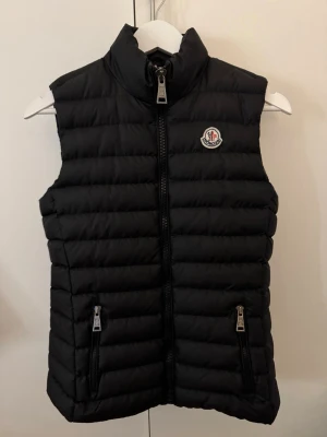 Svart dunväst från Moncler - Säljer en svart dunväst från Moncler med quiltad design och hög krage. Västen har två fickor med dragkedja och ett Moncler-märke på bröstet. Insidan har en cool serietidningsetikett. Perfekt för lager-på-lager och streetstyle.