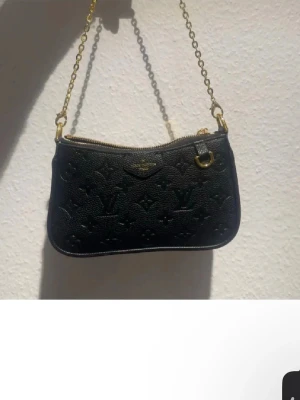 Svart handväska i skinn - Snygg svart handväska  i präglat skinn. Väskan har guldfärgade metalldetaljer, kedjerem och dragkedja upptill. Perfekt storlek för det viktigaste och en riktigt lyxig accessoar.