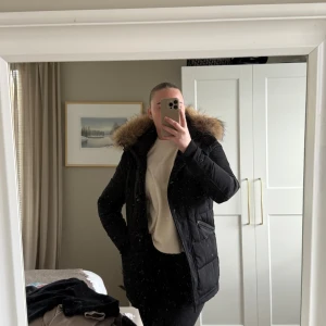 Svart pufferjacka Rockandblue med fuskpäls - Säljer en svart pufferjacka från Rockandblue med stor huva . Jackan har dragkedja, knappar, flera fickor och ribbade muddar vid ärmslut. Perfekt för kalla dagar och riktigt snygg passform.