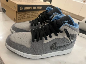 Air Jordan 1 Mid SE Grey Fog/Blue - Air Jordan 1 Mid SE i storlek 44 med grått och blått tyg, svarta snören och detaljer. Swoosh-loggan har en unik broderad look och sulan är tillverkad av återvunnet material med prickigt mönster. Insidan är fodrad med ljusblå textil för extra komfort.