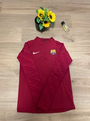 Vinröd Nike FC Barcelona träningströja - Snygg vinröd långärmad träningströja från Nike med FC Barcelona-logga på bröstet. Tröjan har halv dragkedja och diskret mönster på ärmarna. Perfekt för dig som älskar fotboll och vill ha en sportig look.