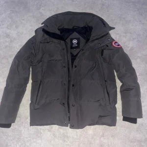 Grå wyndham jacka från Canada Goose - Grå wyndham jacka från Canada Goose med klassisk patch på ärmen, hög krage och flera fickor med dragkedja. Jackan har en quiltad design och är fodrad med mjukt material för extra värme. Perfekt för kalla dagar och har en snygg, stilren look. Canada goose förpacknings påsen finns också med men den svarta väskan har jag tyvärr tappat bort :( en digital kvitto finns!