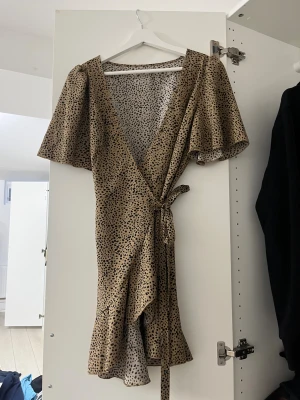 Beige omlottklänning med prickigt mönster - Snygg kort omlottklänning i beige med svart prickigt mönster. Klänningen har v-ringning, volangkant nedtill och knyt i midjan. De korta ärmarna är luftiga och ger en avslappnad vibe. Perfekt för dig som gillar trendiga detaljer och mönster.