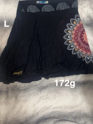 Svart kjol med mandalamönster Desigual - Svart kjol från Desigual i storlek L med färgstarkt mandalamönster i rosa, orange och vitt på sidan. Kjolen har broderad Desigual-logga i gult och är tillverkad i mjuk bomull med elastan för extra komfort. Perfekt för dig som gillar unika detaljer och grafiska prints.🌸 Ena trycket är uttvättad och syns inte så väl. Storlek L