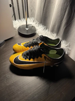 Nike Mercurial fotbollsskor strl 42 - Nike Mercurial fotbollsskor i storlek 42 med en grymt snygg gul och svart randig design. Skorna har snörning, rund tå och platt sula med dobbar för optimalt grepp på planen. Tillverkade i syntetmaterial med vit och svart Nike-logga på sidan. Perfekt för dig som vill sticka ut på fotbollsplanen. Hör av dig vid funderingar och pris kan diskuteras!