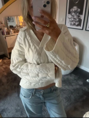 Vit kabelstickad kofta med krage - Säljer en vit kabelstickad kofta med bred krage och v-ringning. Koftan har grova stickade detaljer och ribbade muddar vid ärmslut och nederkant. Modellen är croppad och har en loose passform, perfekt att styla med jeans.