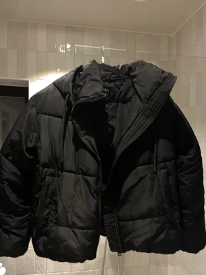 Svart pufferjacka från Zara - Säljer en svart pufferjacka från Zara i storlek S. Jackan har en croppad passform, huva och dragkedja framtill. Den är quiltad och har snörning vid huvan. Perfekt för kalla dagar när du vill vara både varm och snygg.