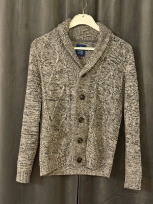 Grå stickad kofta från Tom Tailor - Säljer en gråmelerad stickad kofta från Tom Tailor. Storleken är i M men sitter mer som S. Koftan har tydliga kabelstickade mönster på framsidan, bred sjalkrage och fem knappar framtill. Den är långärmad och har ribbade muddar samt två fickor fram. Skriv vid minsta fundering😁