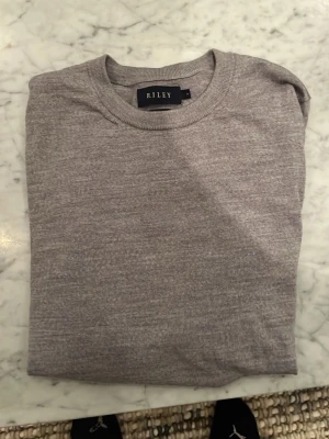 Grå merinoulltröja från Riley - Snygg grå tröja från Riley i mjuk merinoull. Klassisk rund hals och långärmad modell med normal passform. Perfekt att bära till jeans eller chinos för en clean look. Materialet är tunt och skönt mot huden, och färgen är diskret och lättmatchad. Litet hål på höger ärmen
