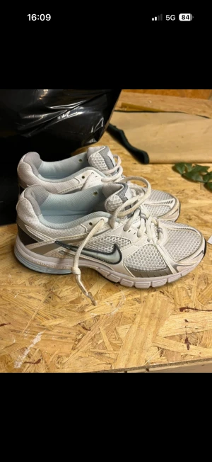 Nike sneakers vita och grå mesh - Nike sneakers i vit och grå färg med mesh-överdel och silvriga detaljer. Snygg svart swoosh på sidan, platt sula och snörning. Skorna har sportig vibe och är perfekta för dig som gillar retro löparstil. Insidan har röd Nike+ logga. Runda tår och ventilerande material.
