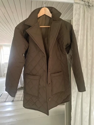 Quiltad brun rock med fickor - Säljer en olivgrön quiltad rock med stora framfickor och knappar. Jackan har en klassisk krage och långärmade ärmar. Perfekt för dig som gillar stilren och enkel design med lite edge. Passar dig som vill ha en jacka med lite längre modell.