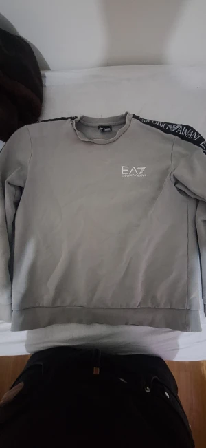 Grå EA7 Emporio Armani sweatshirt - Snygg grå sweatshirt från EA7 Emporio Armani med logga på bröstet och svart band med vit text längs axlarna. Tröjan har rund hals och långa ärmar, insidan är mjuk och fleece-liknande. Perfekt för chill eller streetwear.