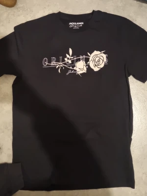 Svart t-shirt med rosprint Jack & Jones - Svart t-shirt från Jack & Jones Originals med vit tryckt text 'ORIGINS' och stiliserade rosor på bröstet. Klassisk rund hals och korta ärmar. Materialet är mjuk bomull som känns skönt mot huden. Perfekt för dig som gillar snygga prints och streetstyle.