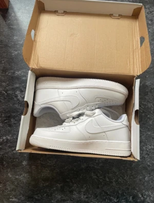 Nike Air Force 1 vita sneakers - Fräscha Nike Air Force 1 sneakers i helvitt skinn med klassisk siluett och låg profil. Skorna har perforerad tå, snörning och platt sula med tydlig Nike-logga på sidan. Perfekta för dig som gillar clean och tidlös streetstil.