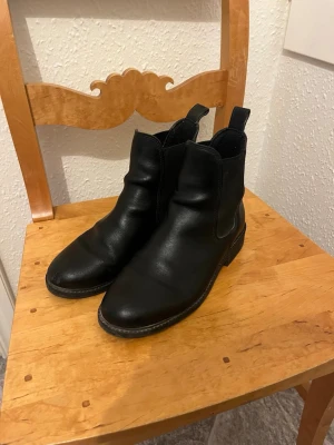 Svarta chelsea boots i skinn - Säljer ett par klassiska svarta chelsea boots i skinn med rund tå och låg klack. Skorna har elastiska paneler på sidorna och dragflikar både fram och bak för enkel påtagning. Perfekta till höst- och vinterlooken.