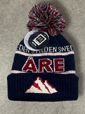 Mörkblå mössa med tofs från Åre - Säljer en mörkblå ribbstickad mössa med stor tofs i rött, vitt och blått. Mössan har texten 'ÅRE' och 'SWEDEN' samt ett broderat bergmotiv framtill. Sprillans nya finns endast att köpa i Åre på plats!