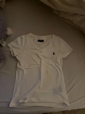 Vit v-ringad t-shirt från Ralph Lauren - Vit t-shirt från Ralph Lauren med klassisk v-ringning och korta ärmar. T-shirten har den ikoniska broderade loggan i mörkblått på bröstet. T-shirten har en fläck. Som går bort, inte pallat försöka bara😊därav priset