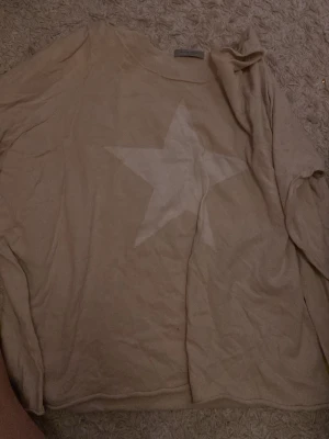 Beige tröja med stjärna från Hunkydory - Säljer en beige långärmad tröja från Hunkydory med en stor vit stjärna framtill. Tröjan är i mjukt material och har en avslappnad passform. Perfekt för dig som gillar enkla men snygga detaljer.