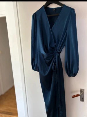 Marinblå omlottklänning med knyt - Snygg marinblå långklänning i satinliknande material med omlottmodell och knyt i midjan. Klänningen har v-ringning och långa puffärmar med resår vid ärmslut. Perfekt för dig som vill ha en elegant och trendig look.